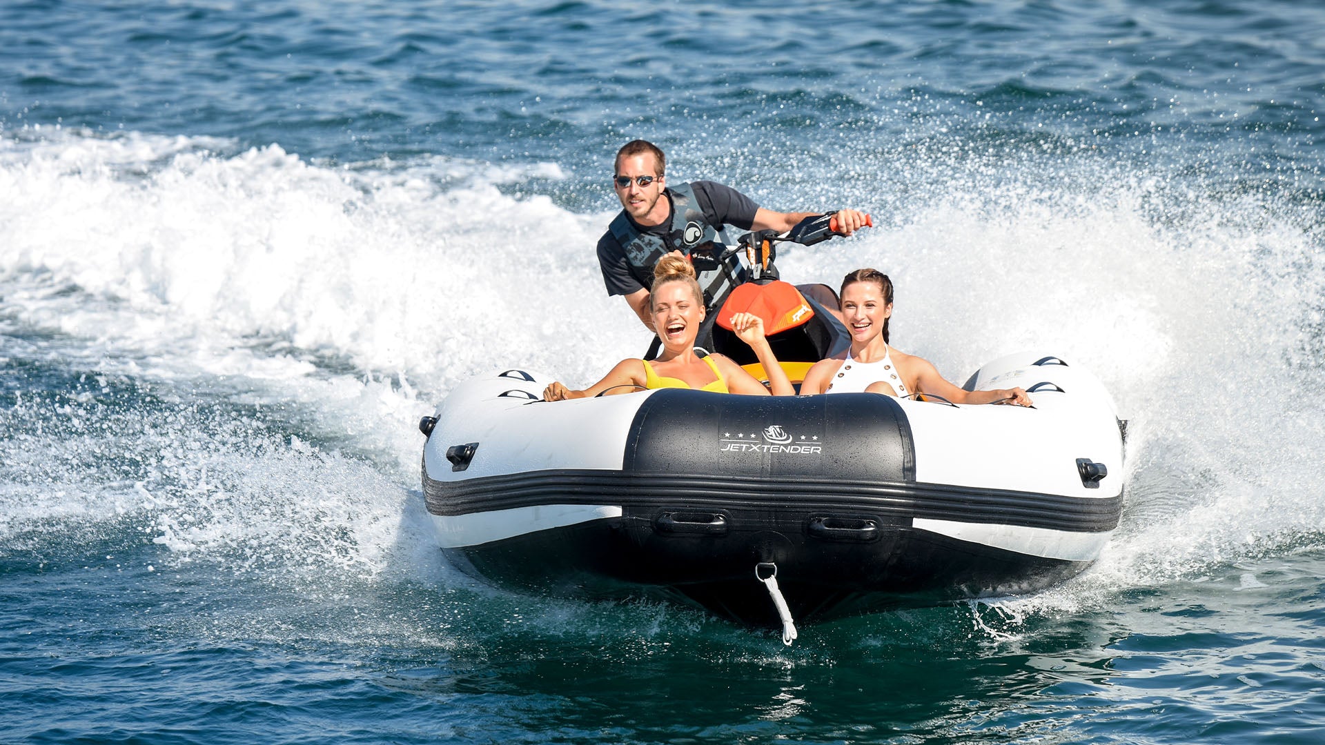 A man is riding a jet ski with the JETXTENDER 380 WELDED attached to the front on which two smiling women are sitting.