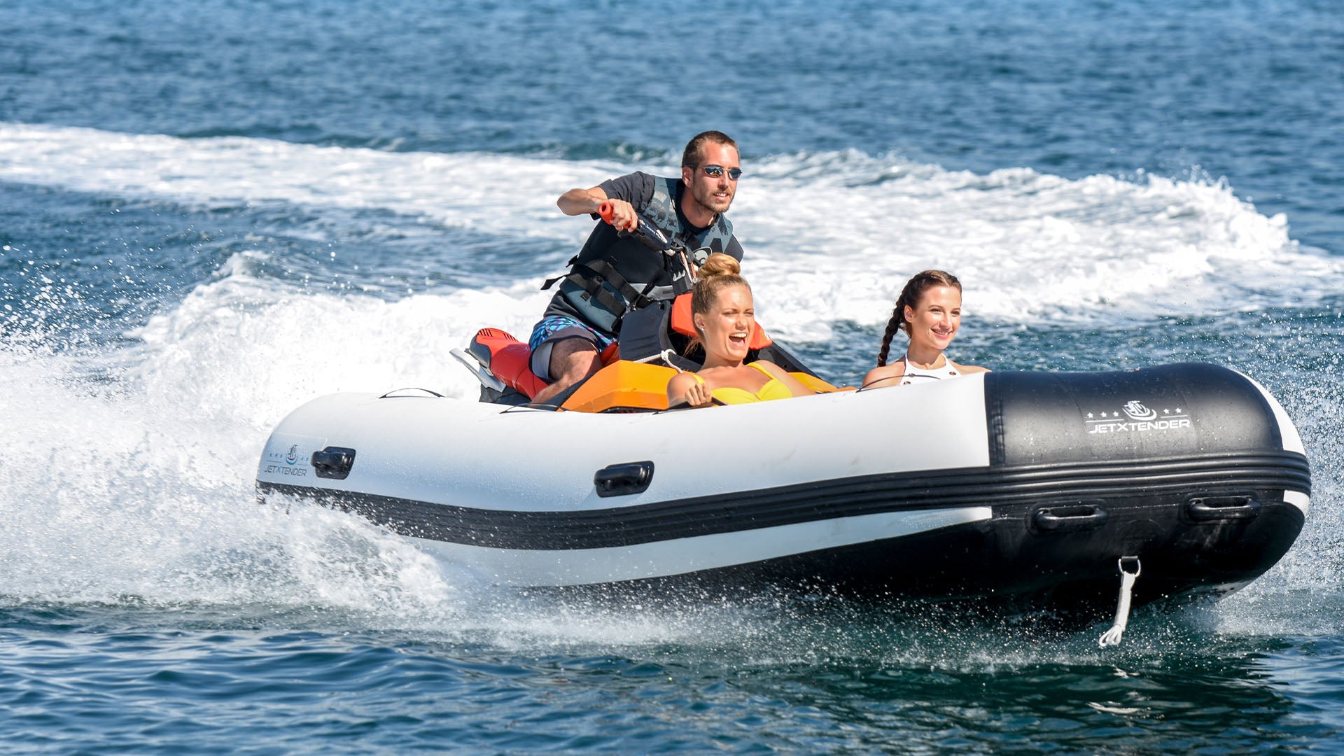 A man is riding a jet ski with the JETXTENDER 380 WELDED attached to the front on which two smiling women are sitting.