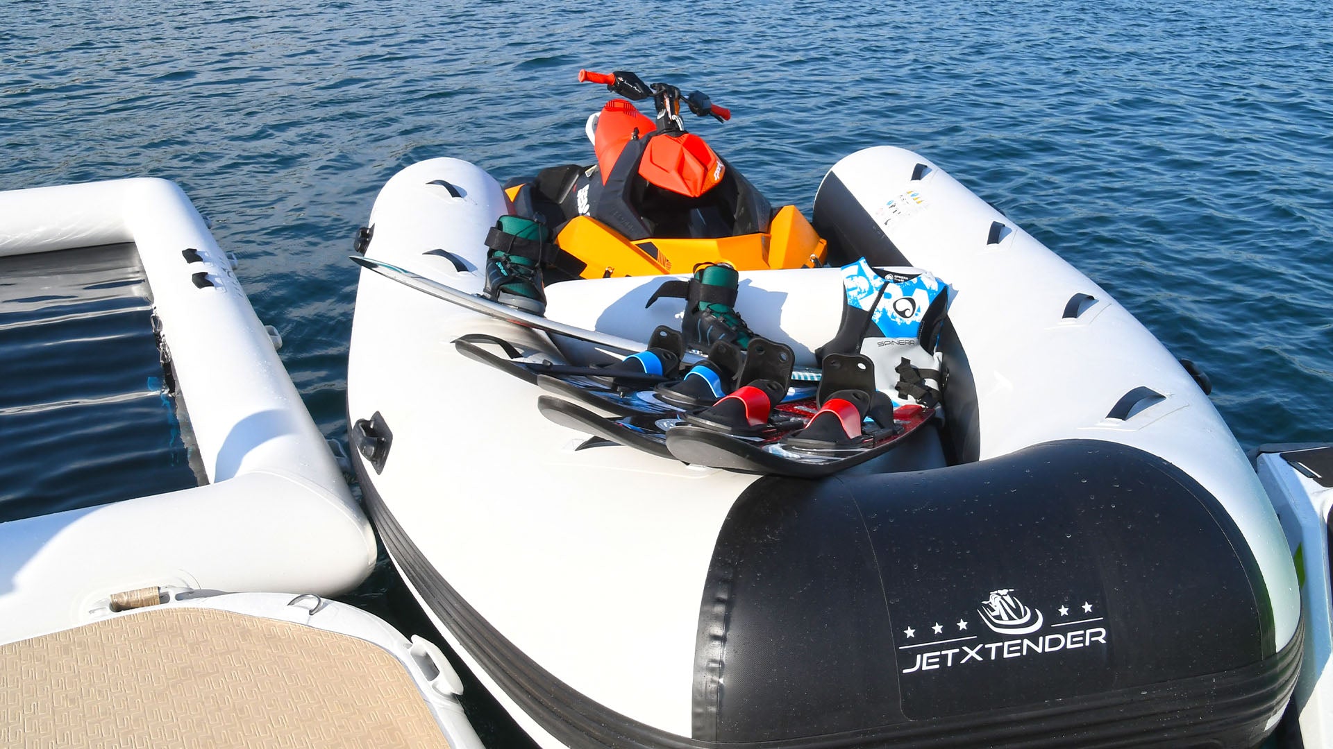 A jet ski is standing next to a boat in the water and has the JETXTENDER attached to the front. Various water sports equipment has been placed on it.