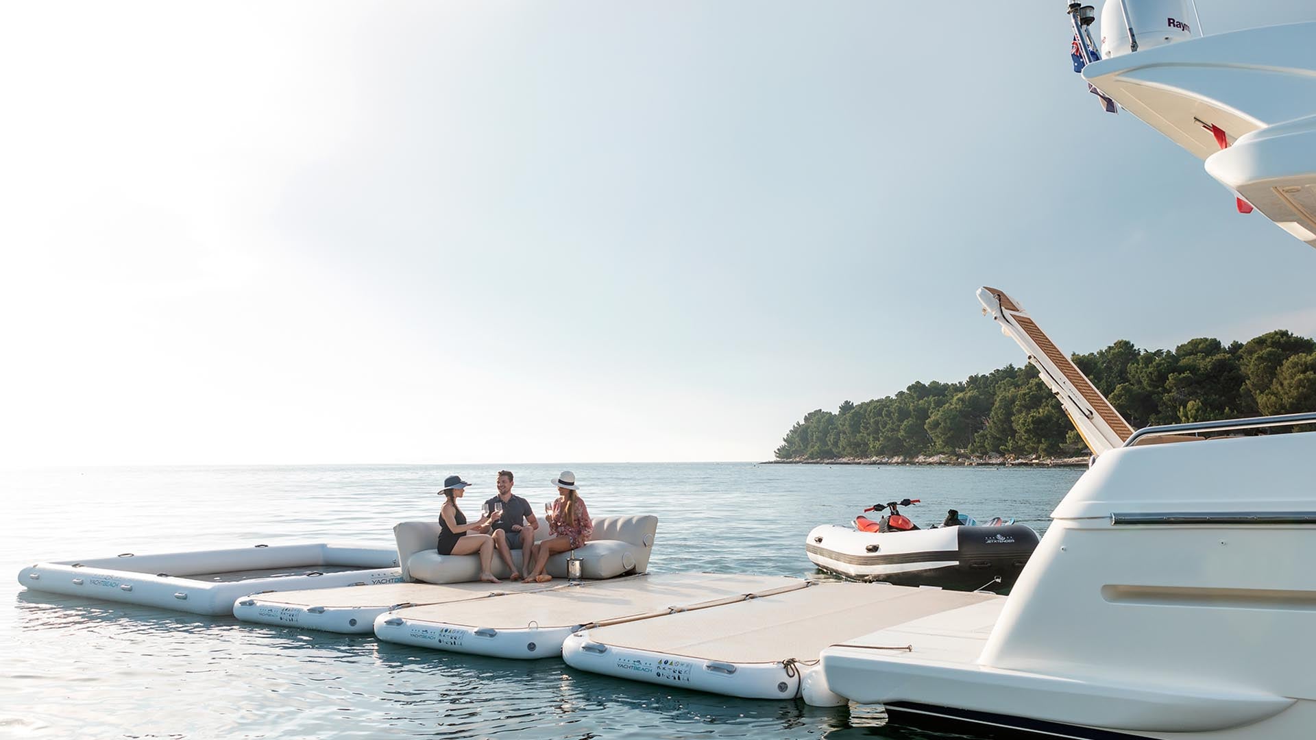 Four yacht beach platforms are attached to a yacht on the water and 3 people sit on them and have a drink.