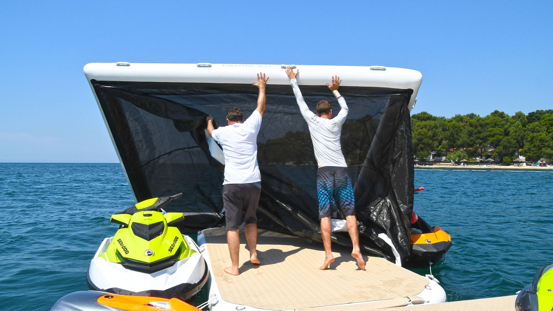 Two men stand on a Yacht Beach platform on the water and set up the Yacht Beach inflatable pool.