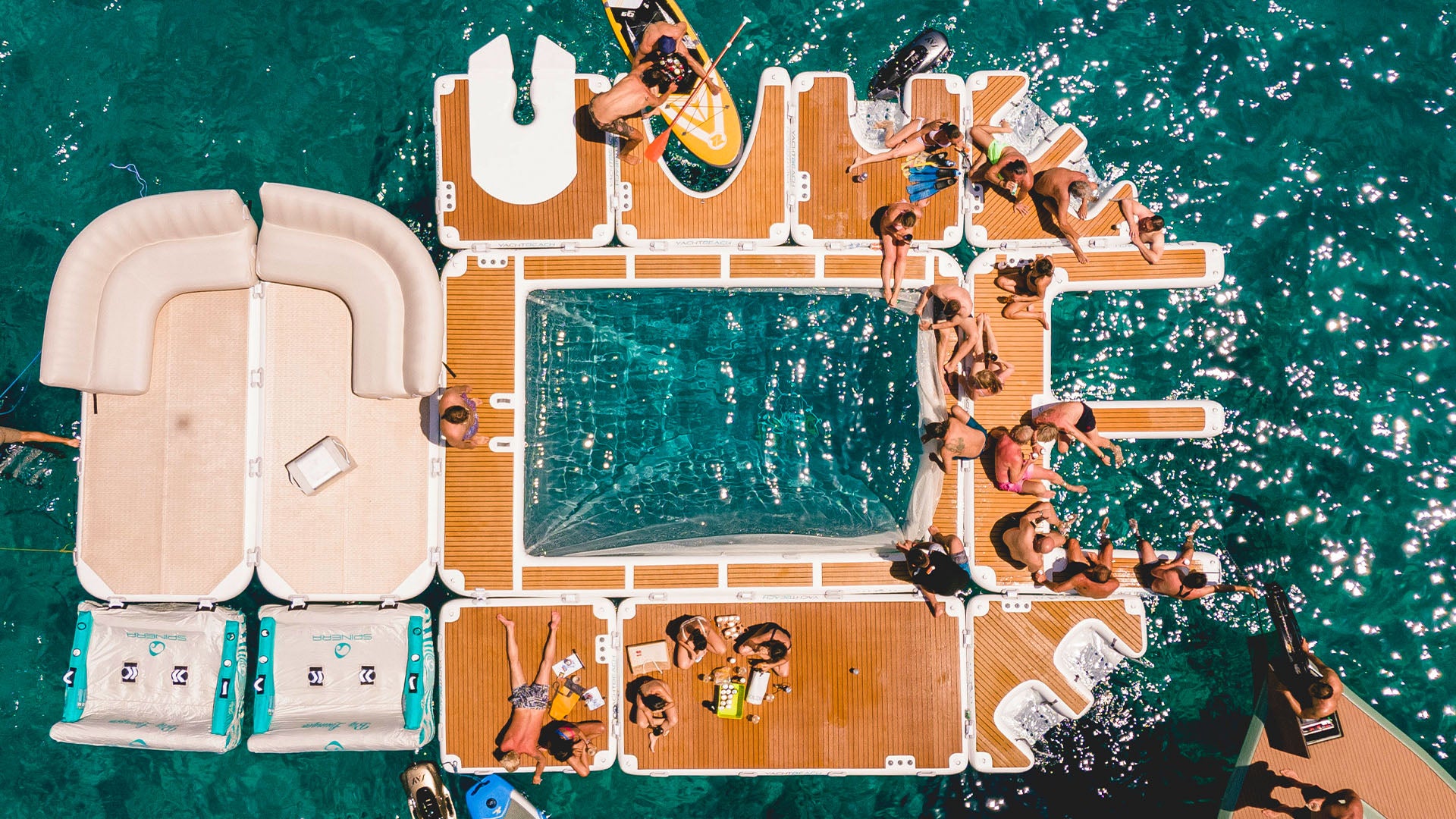 The Yachtbeach luxury pool on the water is linked to other Yachtbeach platforms. Many people have made themselves comfortable on it in their bathing suits.