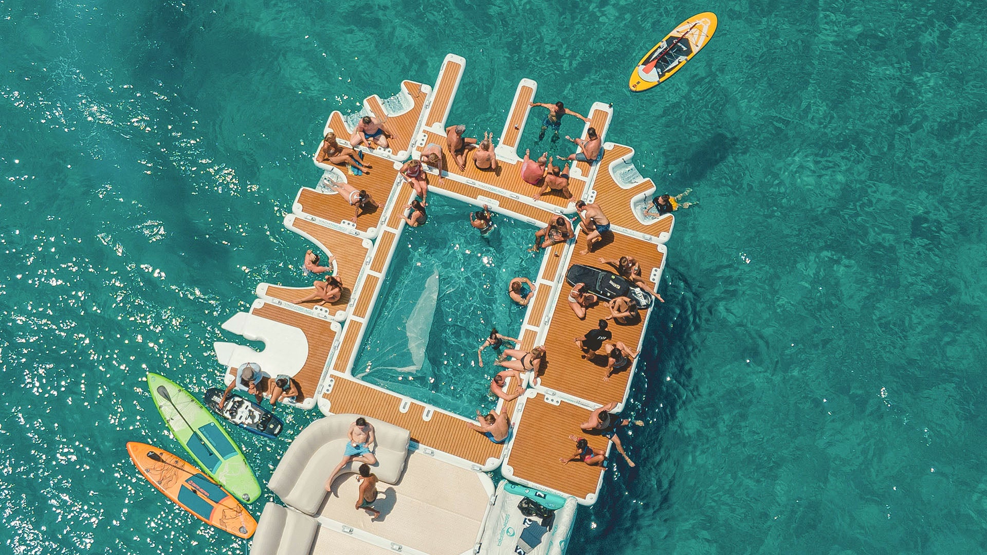 The Yachtbeach luxury pool on the water is linked to other Yachtbeach platforms. Many people have made themselves comfortable on it in their bathing suits.