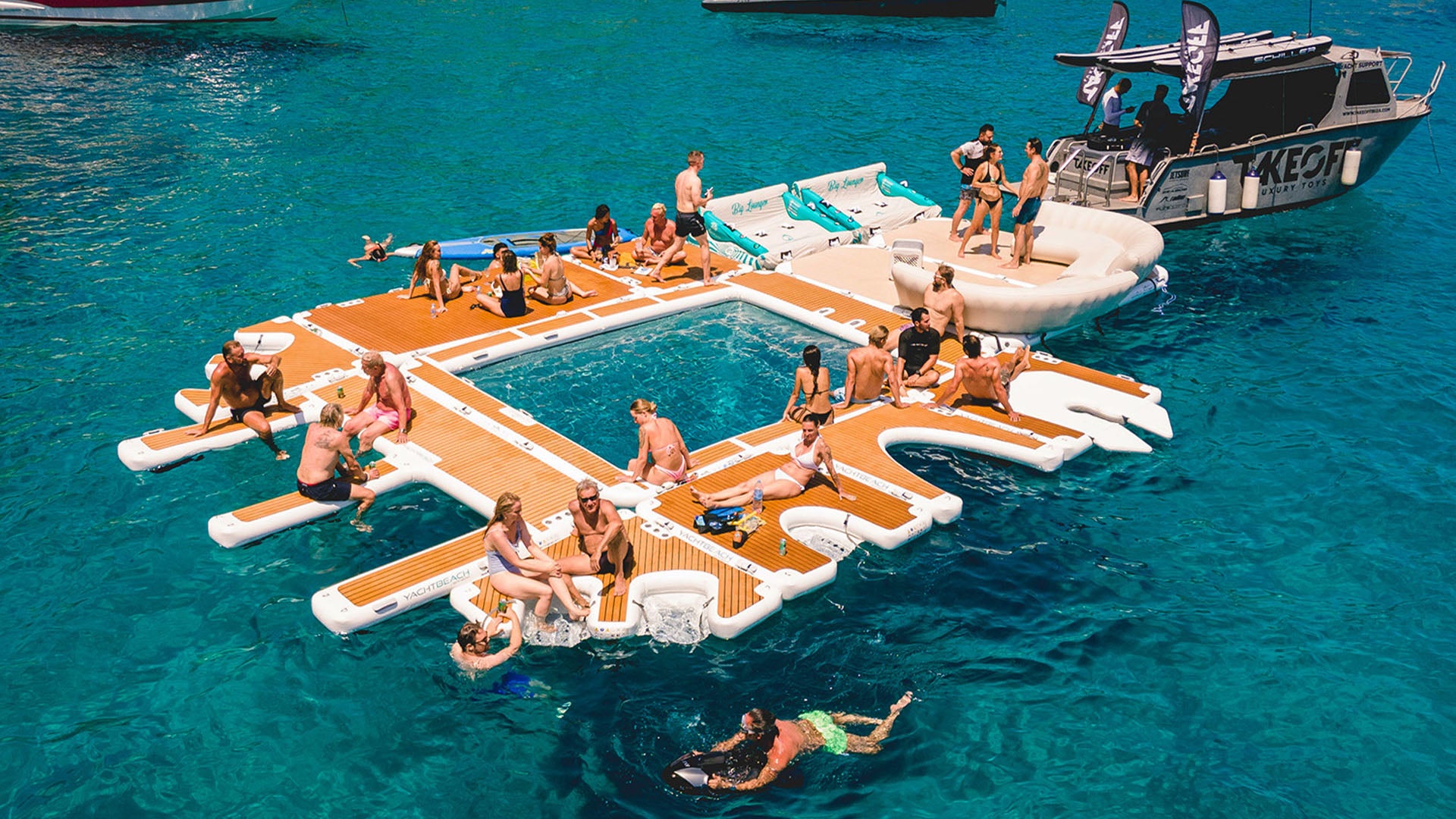 The Yachtbeach luxury pool on the water is linked to other Yachtbeach platforms. Many people have made themselves comfortable on it in their bathing suits.