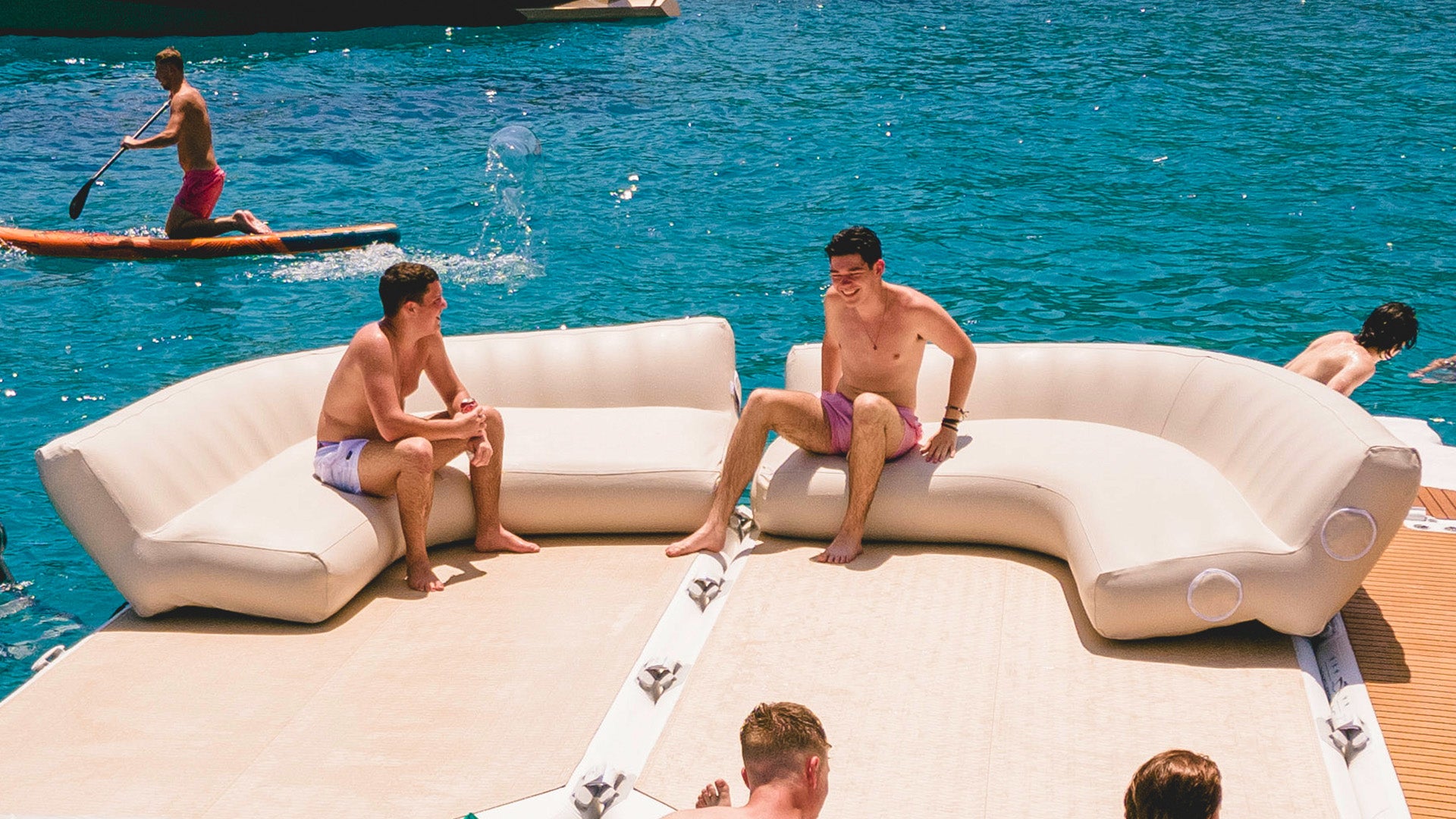 People are sitting in wetsuits on the Yacht Beach Air Sofa on a platform that is floating on the water.