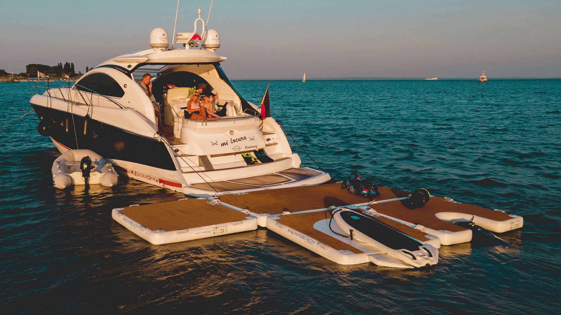 Un barco en el agua tiene varias plataformas flotantes fijadas a la parte trasera, entre ellas un Yacht Beach Foil Dock con un eFoil fijado a él.